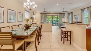 A spacious dining area with a long table, chairs, and a chandelier. There is a kitchen area with a bar and seating. - Games Room | Private Pool with Built in BBQ | 8 Miles to Disney (Kissimmee)