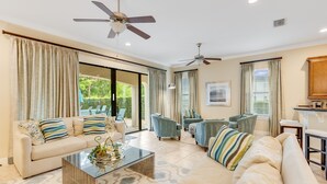 A spacious living room with a beige sofa, armchairs, and a coffee table. There are ceiling fans, large windows with curtains, and a view of greenery outside. - Games Room | Private Pool with Built in BBQ | 8 Miles to Disney (Kissimmee)