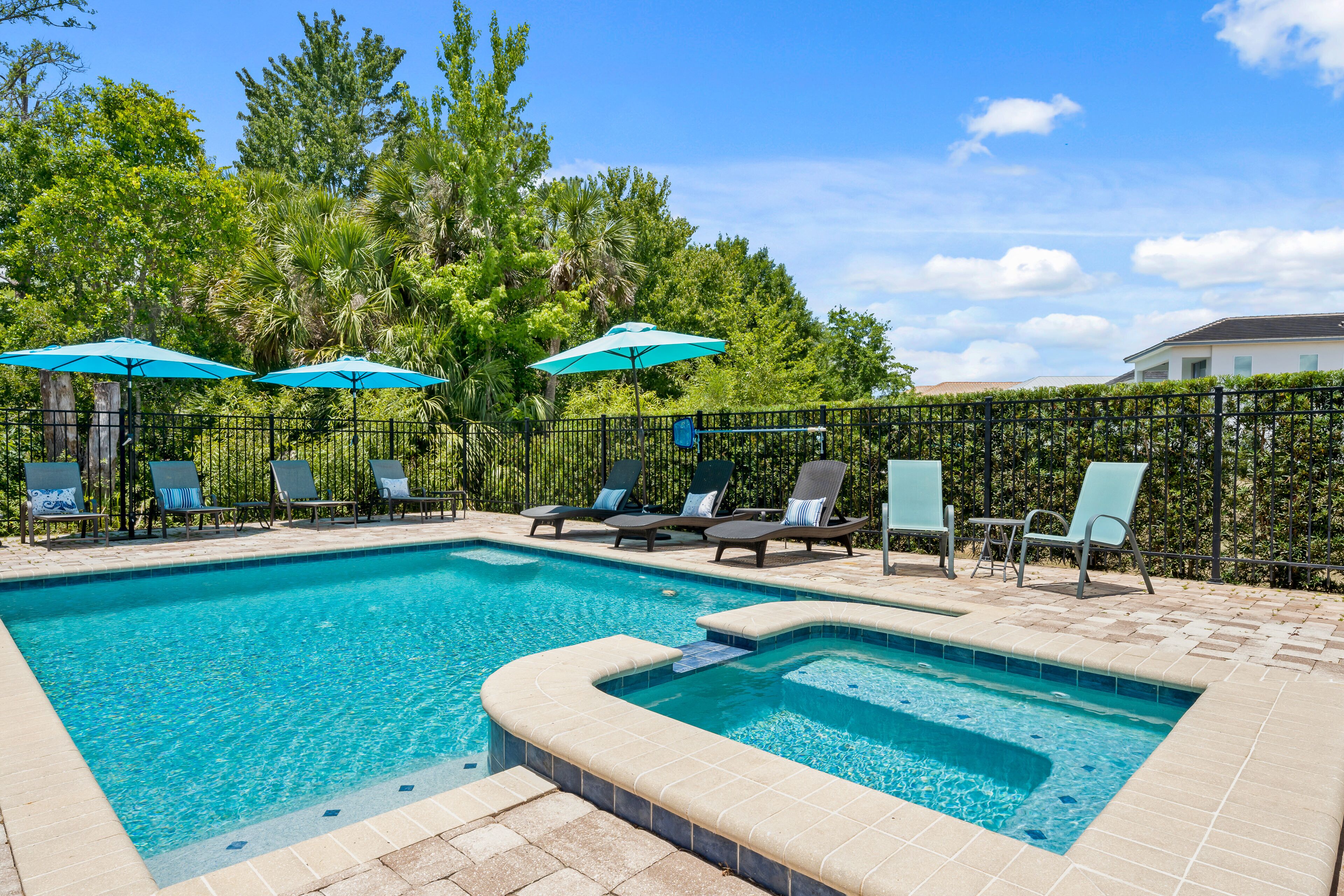 A rectangular swimming pool with a hot tub, surrounded by lounge chairs and umbrellas, set against a backdrop of trees and a clear sky.