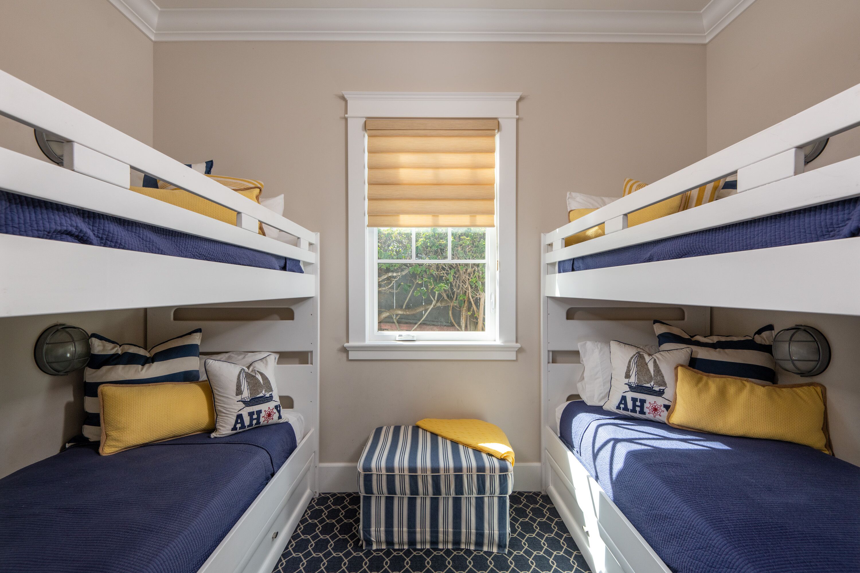 A room with two bunk beds, a window with blinds, and a small ottoman between the beds.
