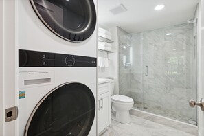 A modern bathroom with a glass-enclosed shower, a white toilet, and a wall-mounted washing machine. - Seas the Day - Stepless -Walking distance from beach - New remodel interior 2023 (Hilton Head Island)