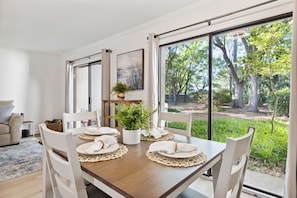 A dining area with a wooden table set for four, white chairs, and a view of a garden through large windows. - Seas the Day - Stepless -Walking distance from beach - New remodel interior 2023 (Hilton Head Island)