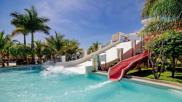 Una piscina con un tobogán rojo, rodeada de palmeras y un edificio con techo inclinado.