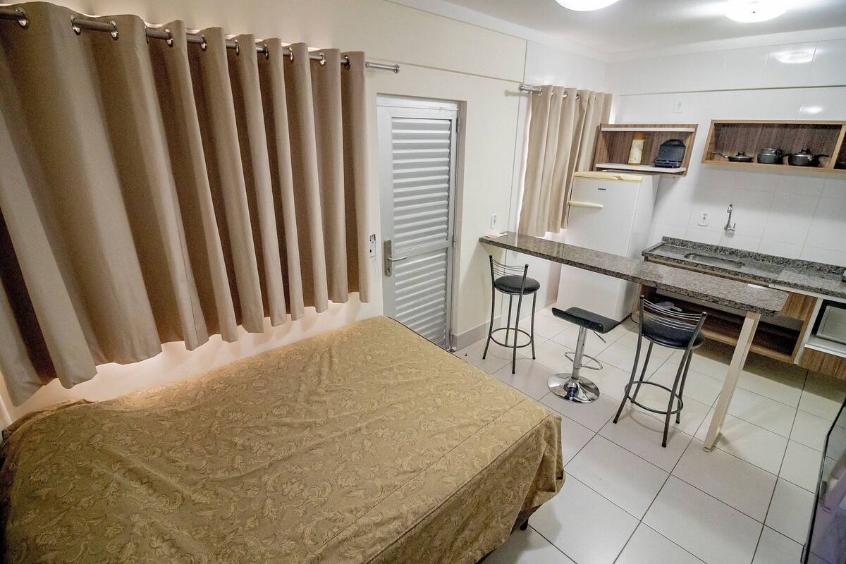 A compact hotel room with a bed, a small kitchenette, and a dining area with two stools.