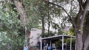 Un espace extérieur aménagé, abrité et doté de sièges, avec une balançoire, le tout entouré d’arbres et d’une pelouse.
