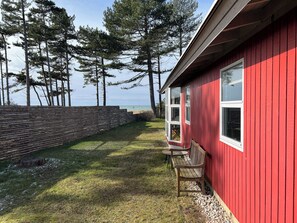 A red building with a bench and a table outside, overlooking a grassy area and trees. - Holiday home on the beach with panoramic view (Store Fuglede)