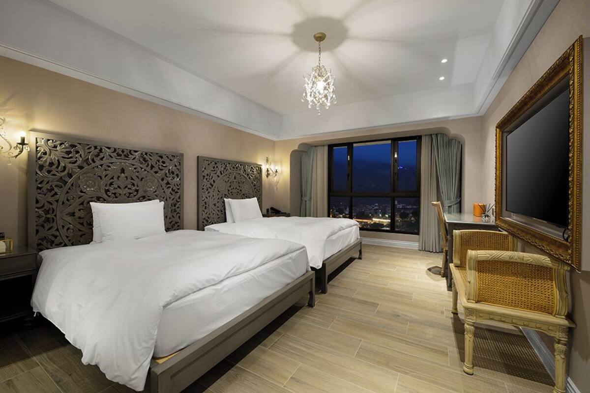 Camera d'albergo con due letti, un lampadario, un'ampia finestra vista città e un televisore.