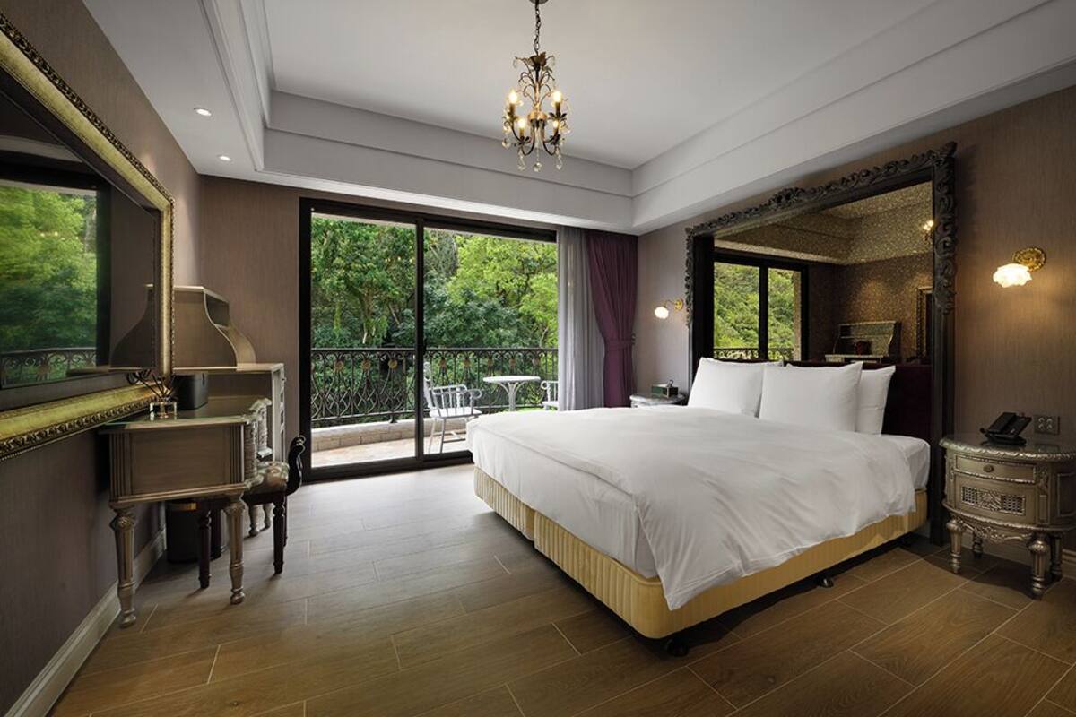 Un'ampia camera da letto con un letto grande, un lampadario, un comodino e vista sul verde dalle finestre.