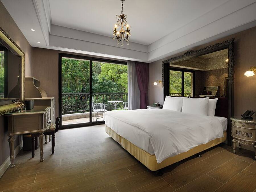 Ein geräumiges Schlafzimmer mit einem großen Bett, einer Kandelaberleuchte, einem Nachttisch und Blick auf Grün hinter den Fenstern.