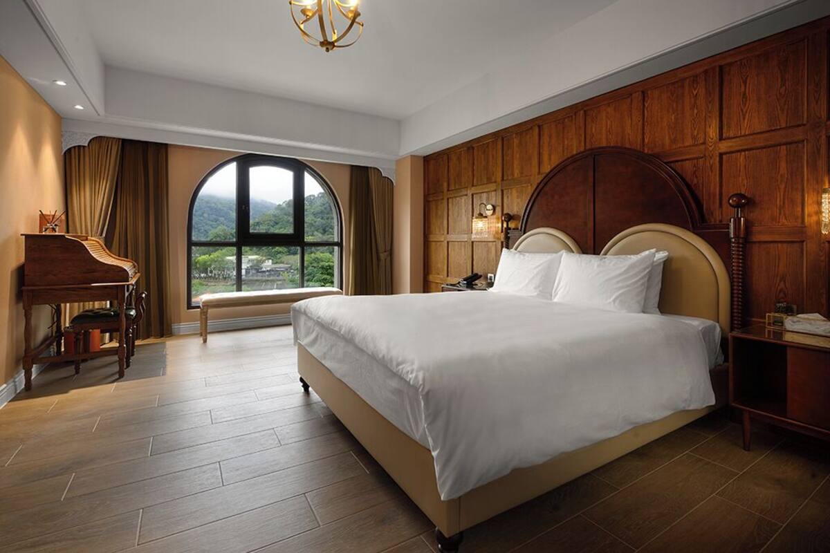 Una camera da letto con un letto grande, pannelli di legno, un pianoforte e vista sul verde attraverso un'ampia finestra.