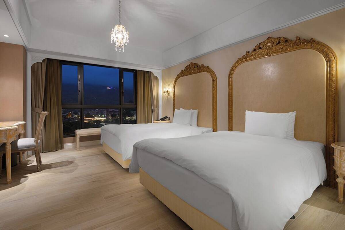 Una camera d'albergo con due letti, una scrivania, una sedia, un'ampia finestra vista città e testiere decorate con cornici dorate.