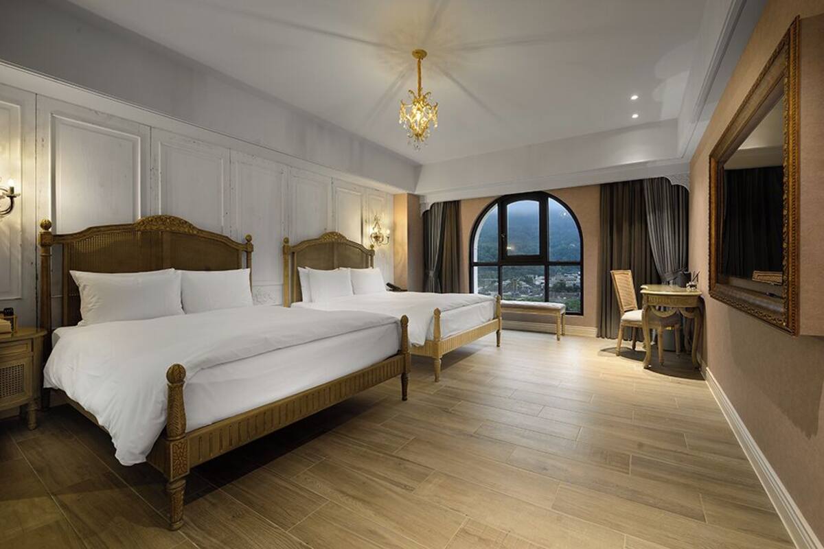 Un'ampia camera da letto con due letti grandi, un lampadario, uno specchio grande e vista sulla città da una grande finestra.