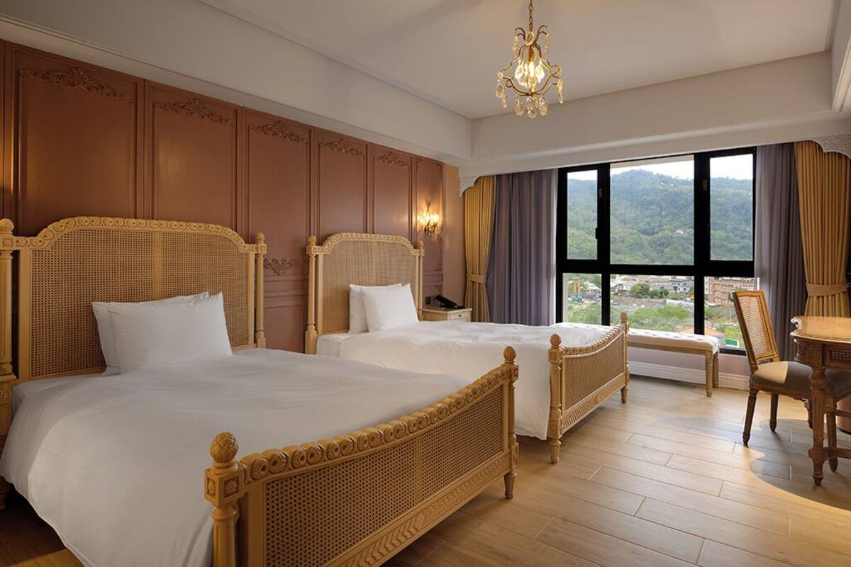 Una camera d'albergo con due letti grandi, un lampadario, vista sull'esterno e un'area pranzo con tavolo e sedie.