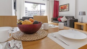 Ein Essbereich mit einem Holztisch, gedeckt für zwei Personen, einer Obstschüssel, weißen Tellern und Besteck. Im Hintergrund befinden sich eine Couch und eine Lampe.
