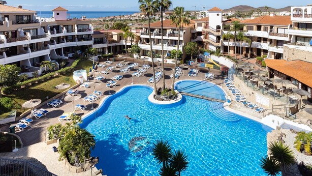 Ein Resort mit einem zentral gelegenen Schwimmbad, umgeben von Liegestühlen und Palmen, mit mehreren Hotelgebäuden im Hintergrund.