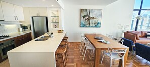 A modern kitchen with a central island, white cabinetry, and a dining area with a wooden table and white chairs. - Penthouse Apartment With Massive Private Terrace + Parking + 2 Pools + Views (Rushcutters Bay)