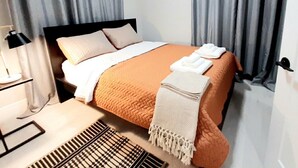 A neatly made bed with a white and orange bedspread, a white throw blanket, and a folded towel on the side. There is a black lamp on a bedside table, a patterned rug on the floor, and grey curtains on the windows. - Cozy, Modern and Centrally Located (Edmonton)