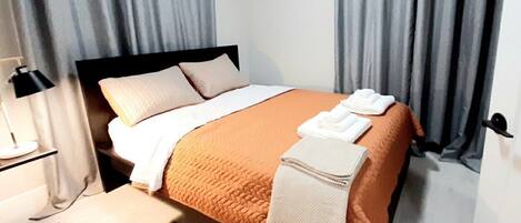 A neatly made bed with a white and orange bedspread, a white throw blanket, and a folded towel on the side. There is a black lamp on a bedside table, a patterned rug on the floor, and grey curtains on the windows.