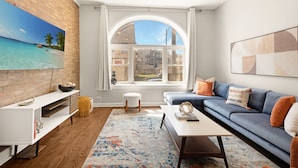 A modern living room with a large window, a white console table, a blue sofa, and a colorful rug. - 4BD Condo w/Parking & Game Room (Chicago)