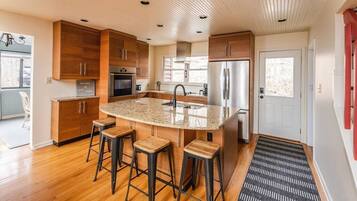 Une cuisine moderne avec des meubles en bois, un îlot central doté de tabourets, des appareils en acier inoxydable et une vue sur l’extérieur.