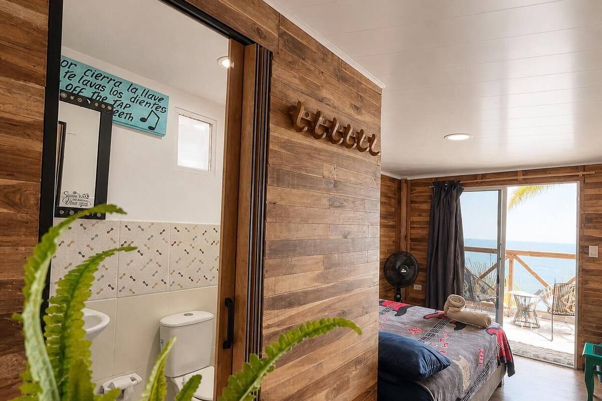 A room with a wooden wall, a bed, a bathroom with a toilet and sink, and a view of the sea.