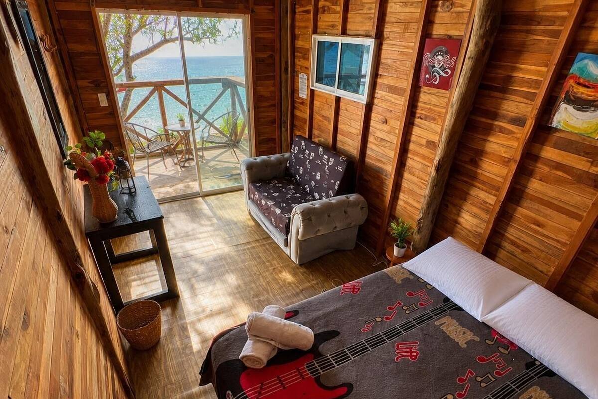 A wooden cabin room with a bed, a chair, a small table, and a view of the sea.