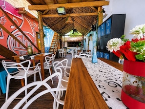 A restaurant with a wooden table, white chairs, a thatched roof, and colorful murals.