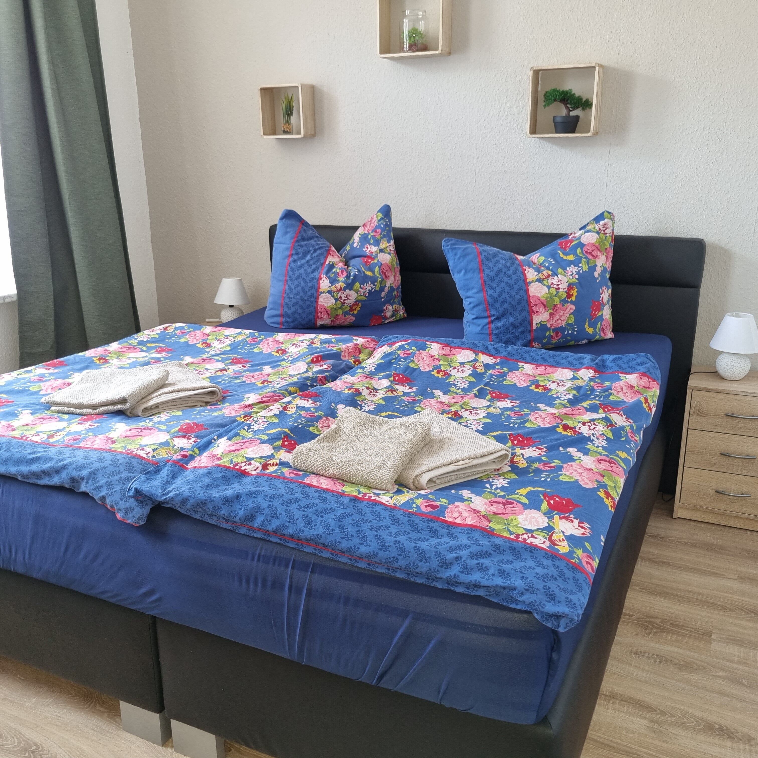 Una cama bien hecha con un edredón floral, almohadas y toallas dobladas. Hay mesitas de noche con lámparas y pequeñas macetas en la pared.
