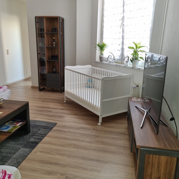 A room with a crib, a wooden cabinet, a small table, and a window with plants.