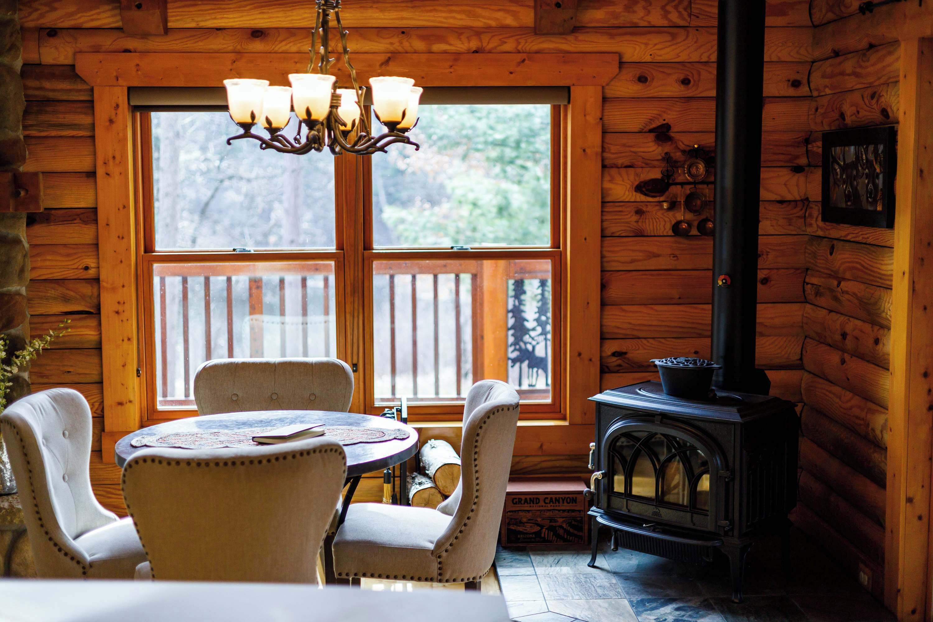A log cabin interior with a dining table, four chairs, a chandelier, a wood-burning stove, and a large window.