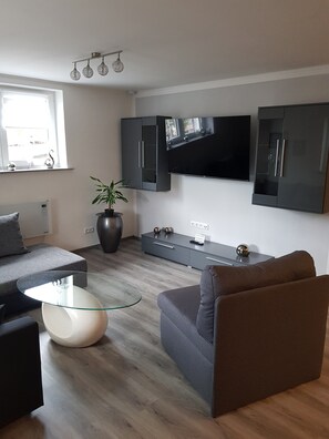 Een moderne woonkamer met een flatscreen televisie, donkere kasten, een glazen salontafel en een potplant.