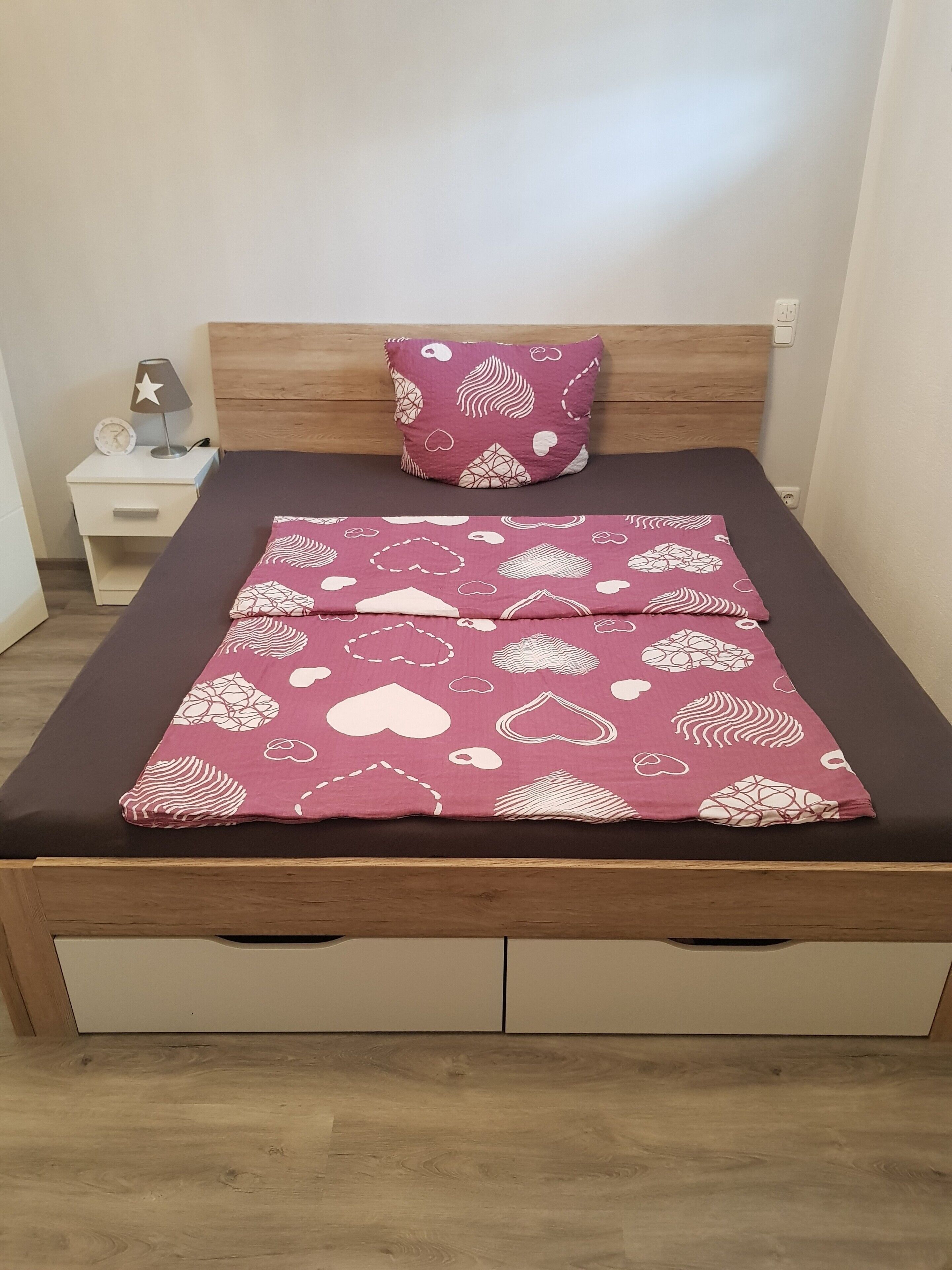A bed with a patterned bedspread, a wooden headboard, a bedside table with a lamp, and a nightstand with a clock.
