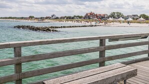 Un muelle de madera con un banco, que conduce a una playa con casas y tiendas de campaña al fondo.