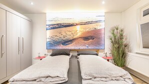 Un dormitorio con una cama grande, una mesita de noche, una planta en maceta y un televisor de pared que muestra una escena de playa.