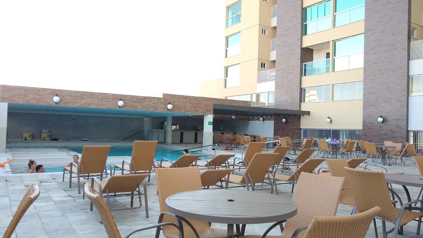 A poolside area with lounge chairs and tables, a pool, and a building in the background.