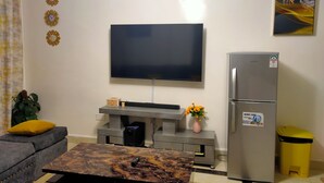 A living room with a flat-screen TV, a grey sofa, a coffee table, a refrigerator, and a trash bin. - 1 Bed Flat Sleeps 2 with On-Site Parking (Nairobi)
