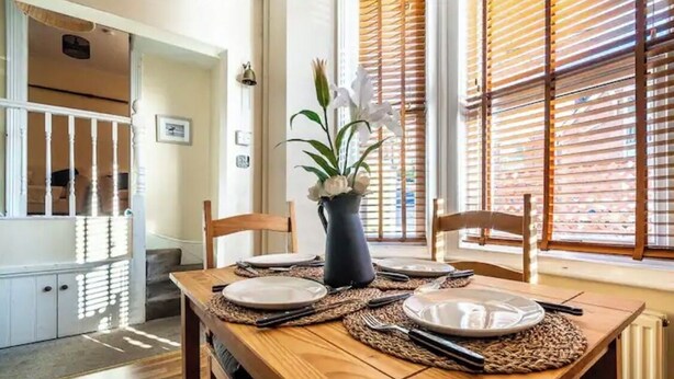 A dining area with a wooden table set for two, a vase with flowers, and a view through wooden blinds. - The Boat House Sleeps 4 Pet Friendly (Dawlish)