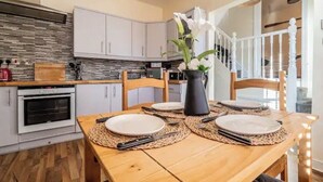 A dining area with a wooden table set for four, a vase with flowers, and a kitchen in the background. - The Boat House Sleeps 4 Pet Friendly (Dawlish)