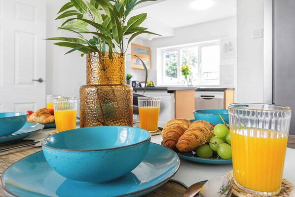 A breakfast setting with a table set for two, featuring blue dishes, a glass of orange juice, croissants, and a green plant in a textured vase.