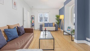 Ein modernes Wohnzimmer mit einem braunen Sofa, einem Glastisch und einem grauen Sofa. Ein Bücherregal mit Büchern und einer Lampe sind ebenfalls vorhanden.