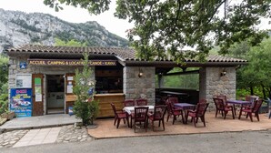 Un restaurante rústico al aire libre con paredes de piedra, mesas y sillas de madera, cerca de una montaña.