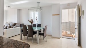 Een moderne eethoek met een glazen tafel en stoelen, een bank met kussens, een kroonluchter en een deur naar een balkon.