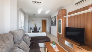 Een moderne woonkamer met een modulaire bank, een flatscreen-tv, houten kasten en een eethoek op de achtergrond.