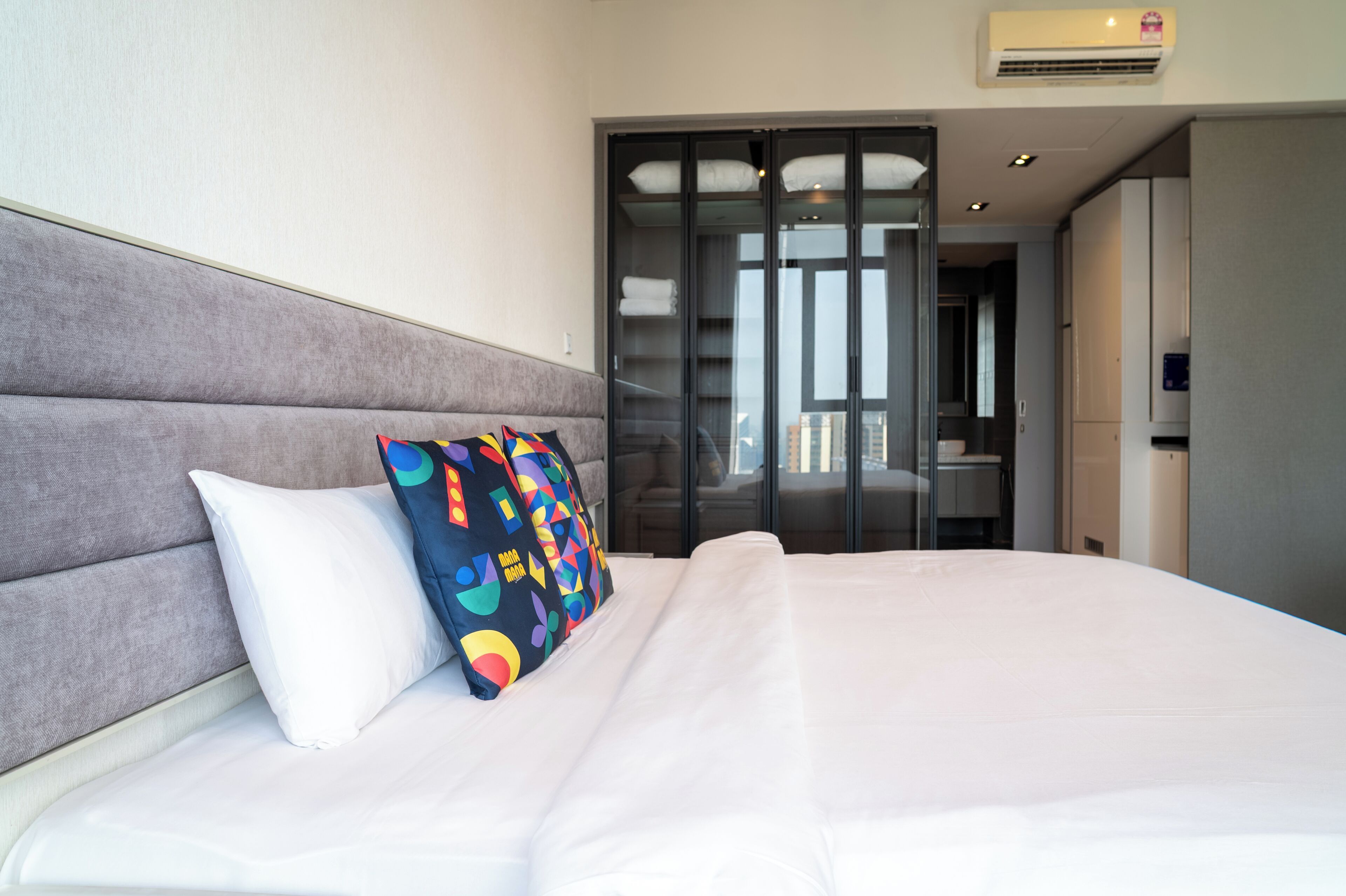 A hotel room with a bed, a grey upholstered headboard, a colorful pillow, and a sliding door closet.