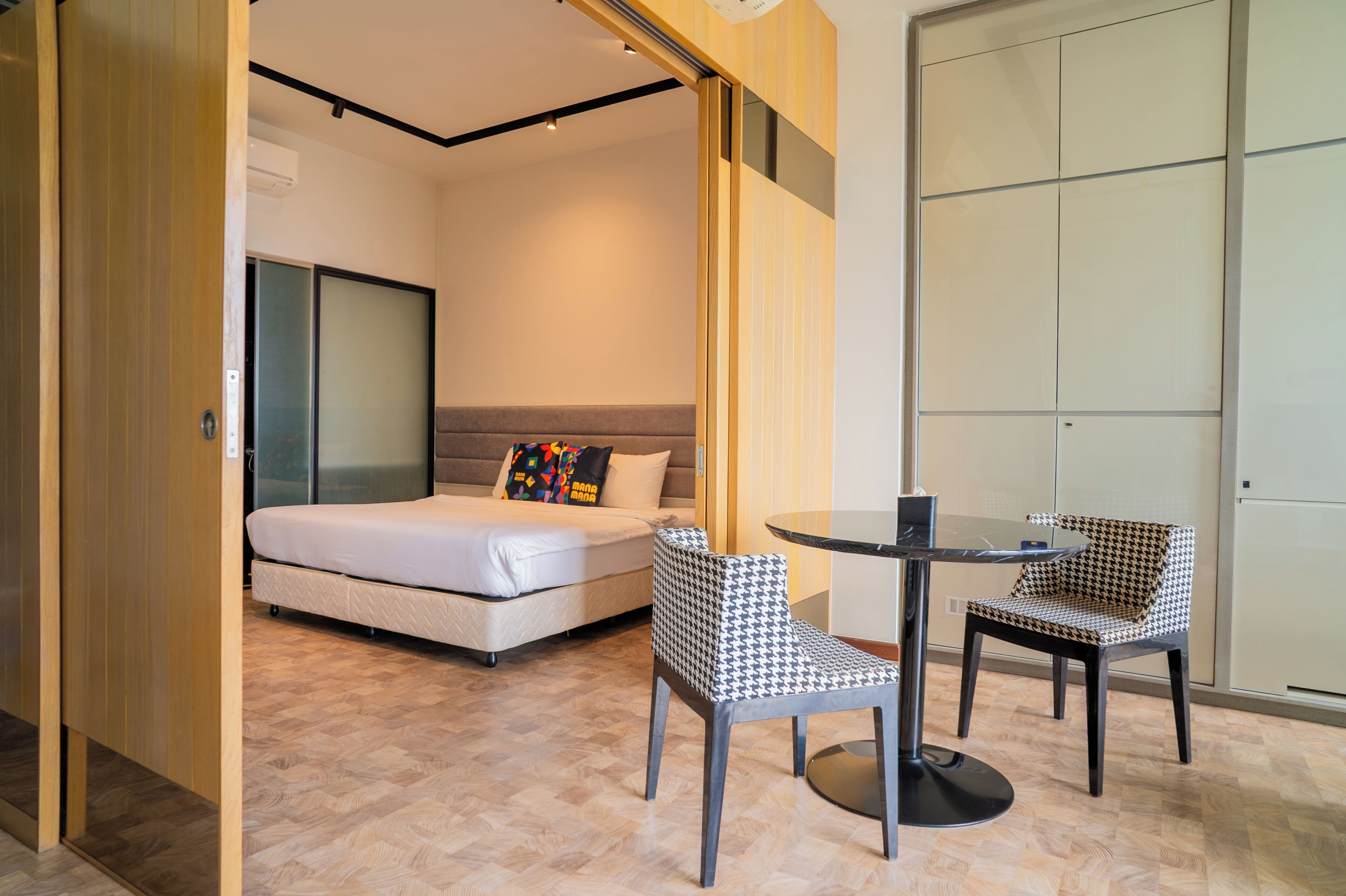 Una habitación de hotel moderna con una cama, dos sillones estampados, una mesita redonda y una puerta corredera que da a otra habitación.