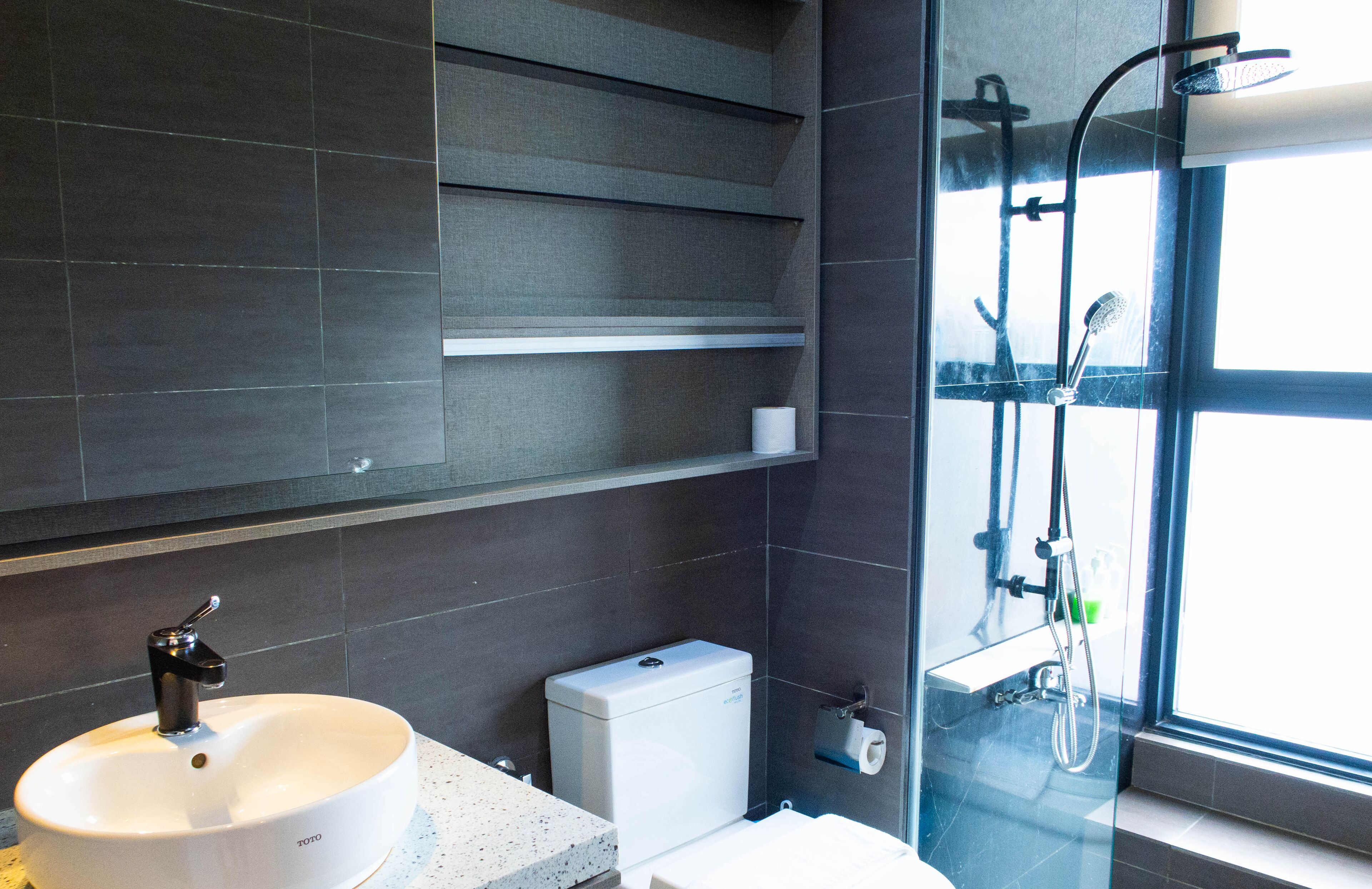 A modern bathroom with a white sink, dark tiled walls, a glass shower enclosure, and a toilet.