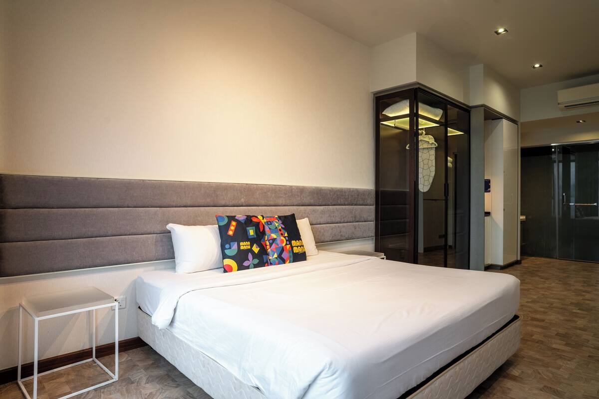 Moderni hotellihuone, jossa on suuri sänky, yöpöytä, vaatekaappi ja lasiovi, joka johtaa kylpyhuoneeseen.