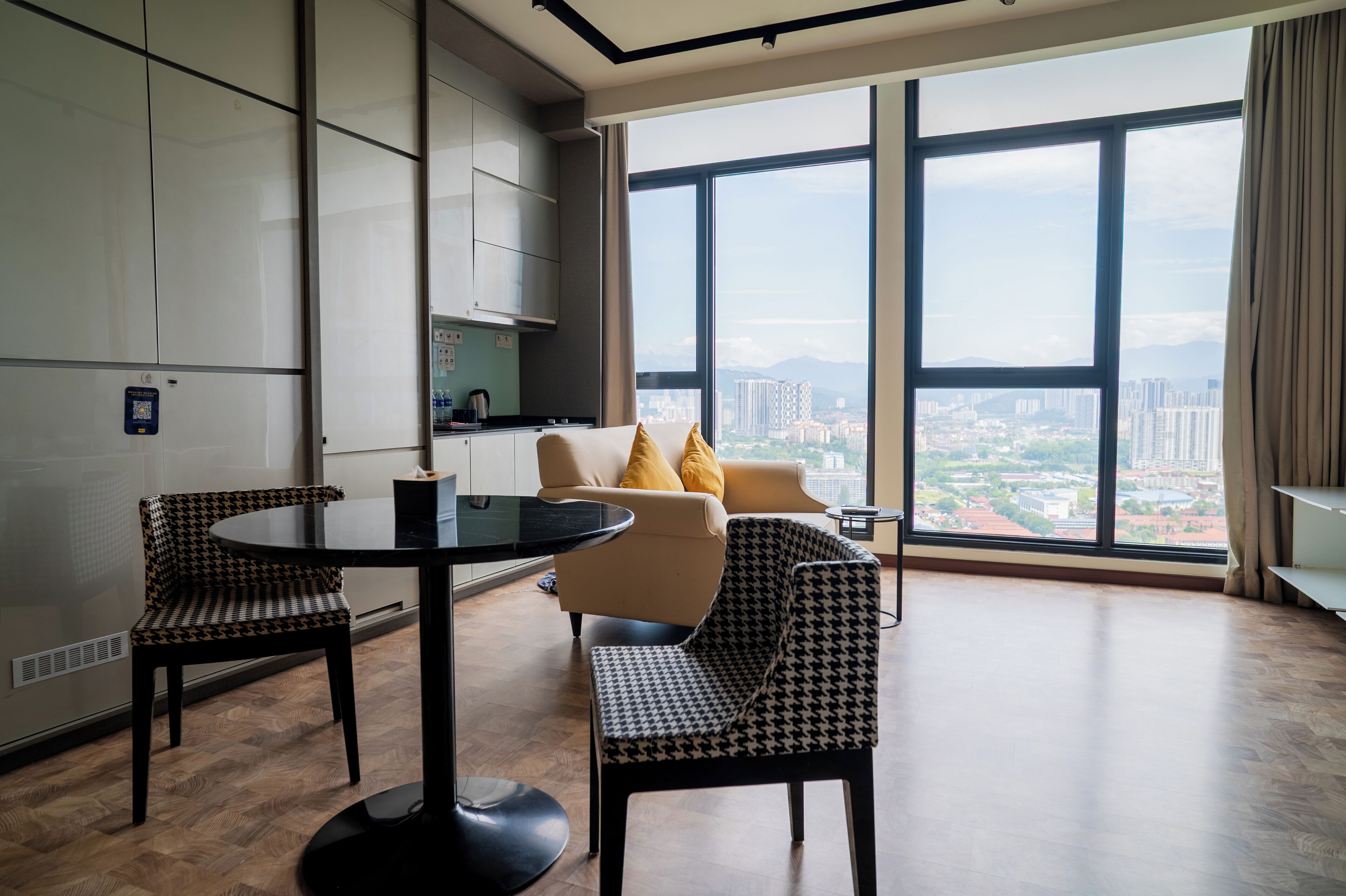 A modern living room with a sofa, two patterned chairs, a round table, and a large window offering a city view.