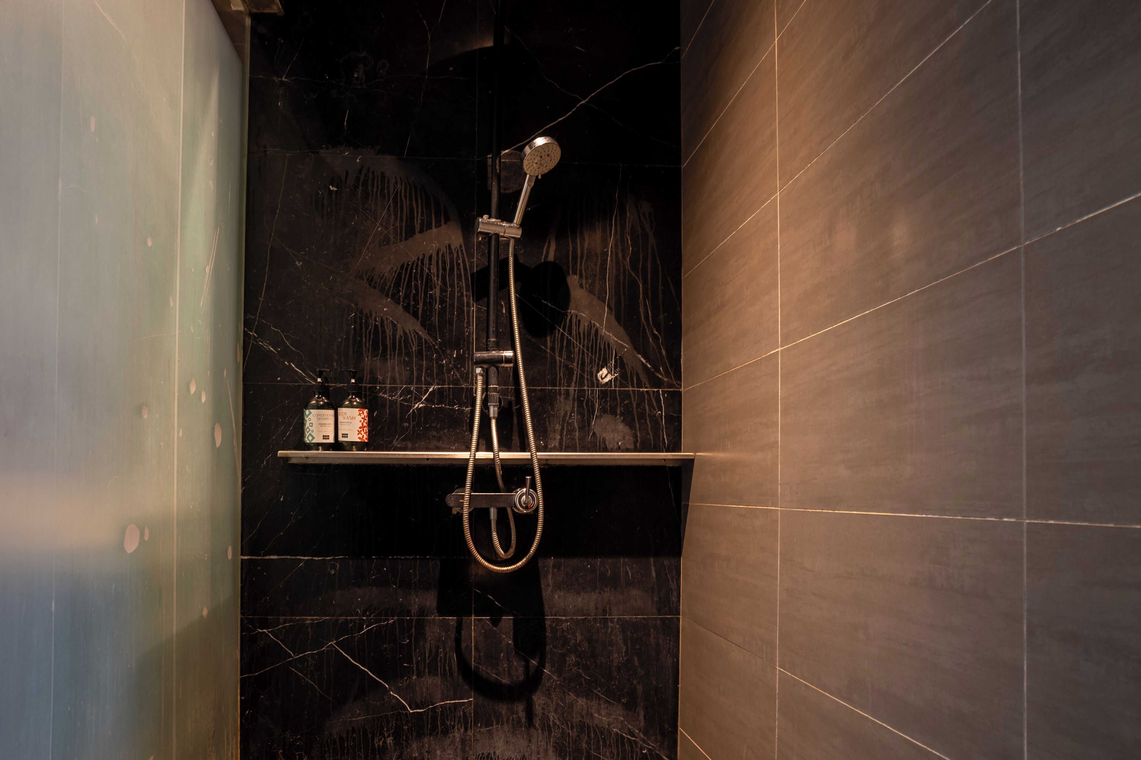 A modern bathroom with a shower, dark marble tiles, and a shelf with toiletries.