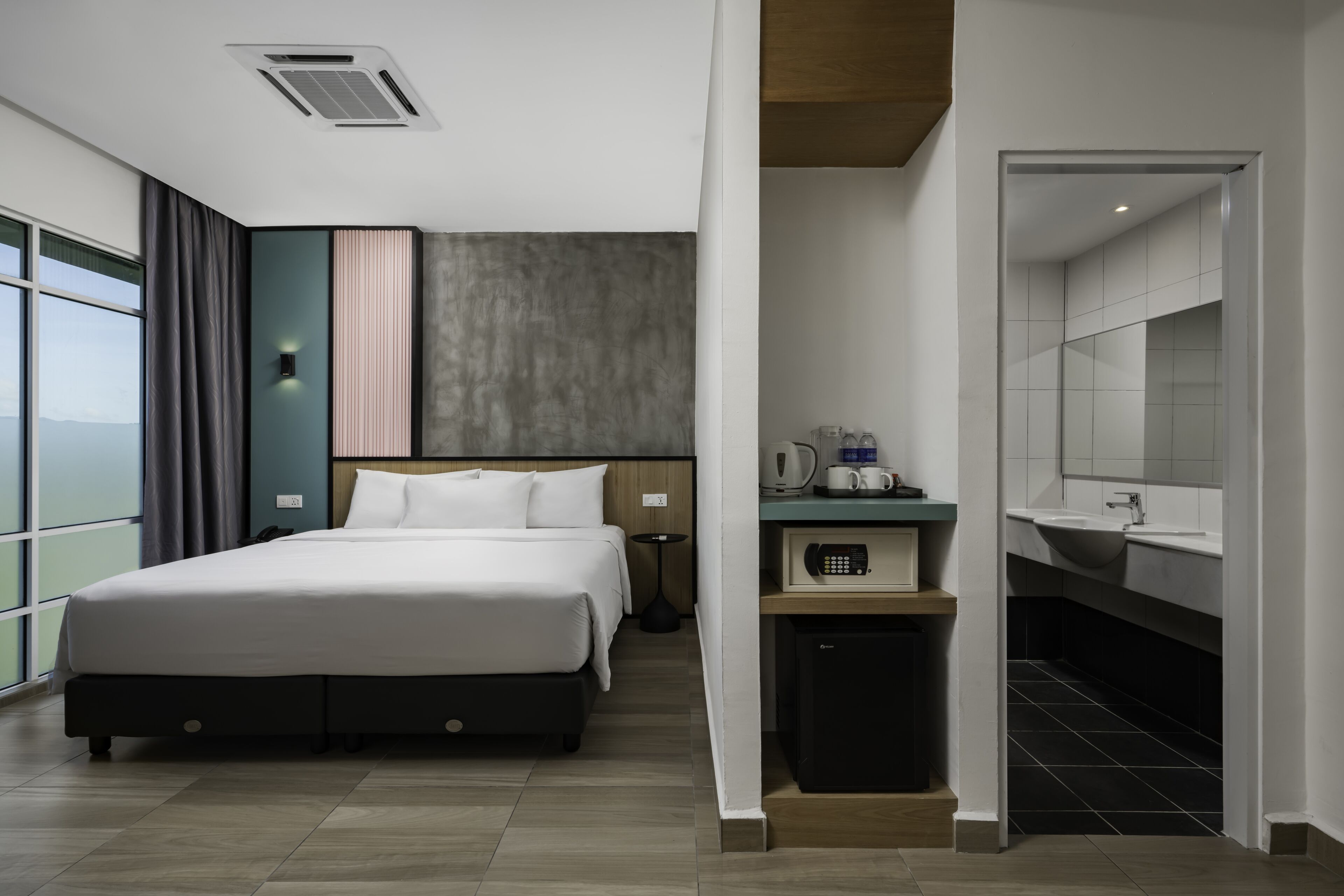 Ein modernes Hotelzimmer mit einem großen Bett, einem Schreibtisch mit Kaffeemaschine, einer kleinen Küchenzeile und einem Badezimmer mit Spiegel und Waschbecken.